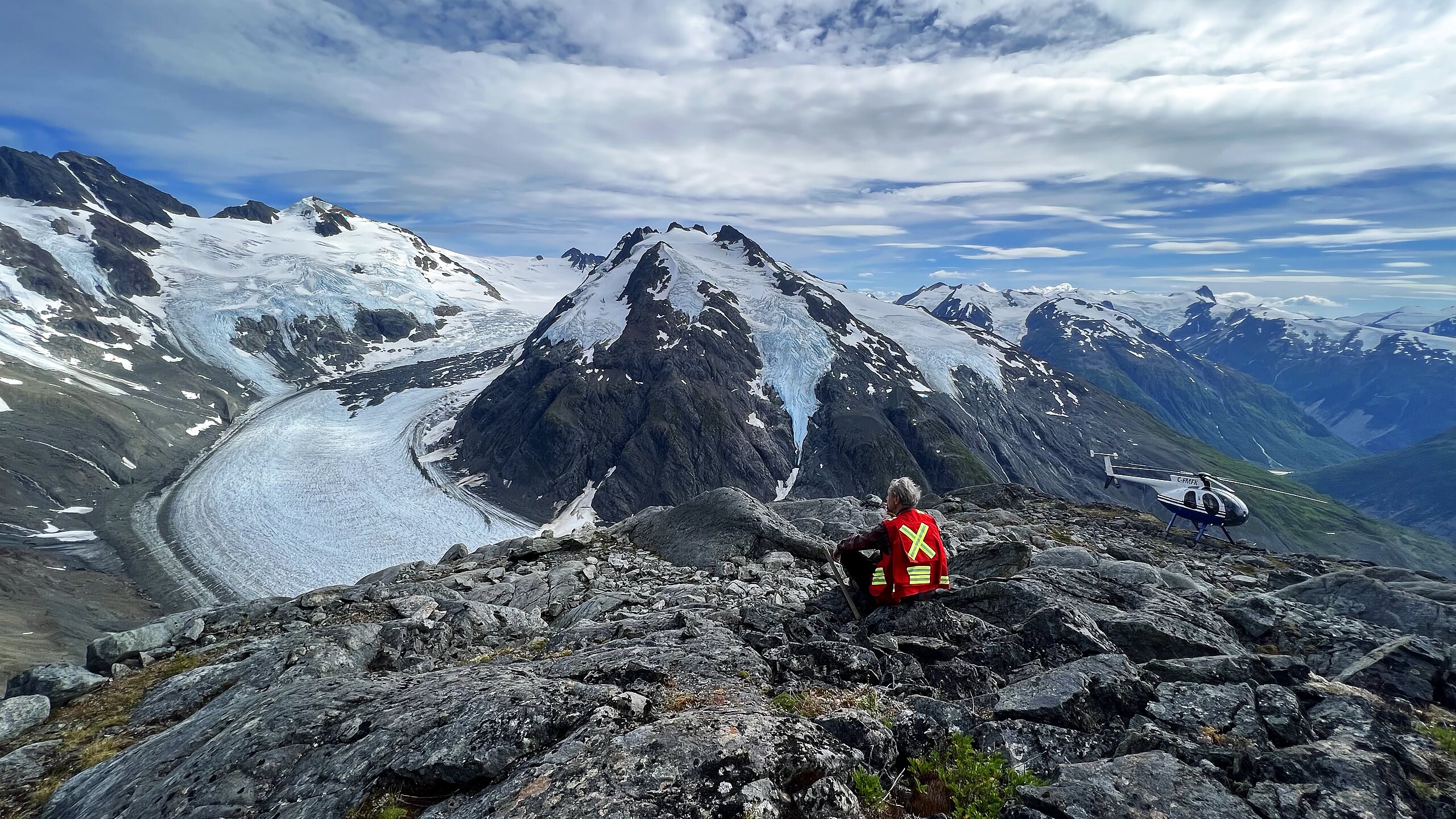 A Luxor Metals field geologist overlooking the Frank Mackie glacier in British Columbia's Golden Triangle, with a support helicopter nearby.
